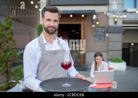 Bärtiger Kellner hält Tablett mit einem Glas Rotwein, eine Kundin sitzt dahinter Stockfoto