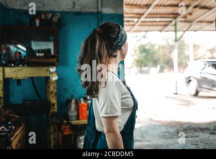 Das Konzept von Kleinunternehmen, Feminismus und Frauengleichheit. Eine junge Frau in Arbeitskleidung schaut von einer Autowerkstatt auf die Straße. Stockfoto