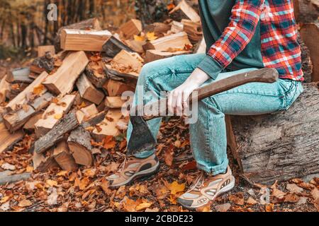Vorbereitung auf die Heizperiode. Eine junge Frau mit einer Axt in den Händen sitzt auf einem Baumstamm. Im Hintergrund ein Haufen Feuerholz. Nahaufnahme. Stockfoto