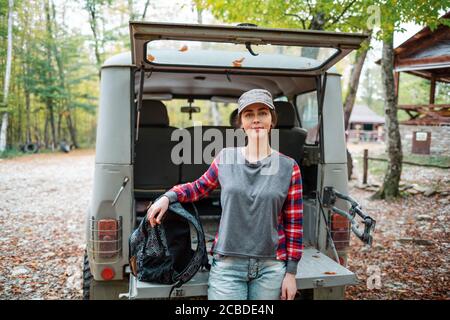 Eine junge kaukasische Frau posiert mit einem Rucksack gegen den offenen Kofferraum eines Jeeps. Im Hintergrund Bäume und Wald. Nahaufnahme. Konzept der Autofahrt A Stockfoto