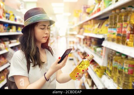 Eine junge schöne Frau hält eine Flasche Öl in der Hand und macht ein Foto von ihr auf einem Handy. Im Hintergrund Regale mit Produkten. Das c Stockfoto