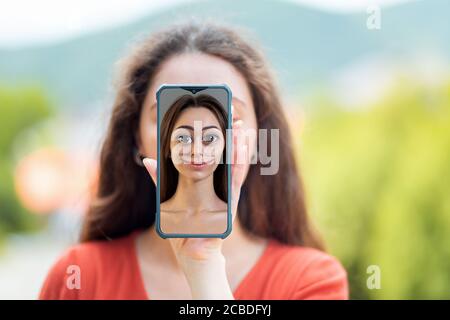 Eine Frau bedeckt ihr Gesicht mit einem Smartphone mit einem Cartoon-Porträt einer Frau. Das Konzept, Identität und gefälschte Seiten in sozialen Netzwerken zu verbergen. Stockfoto