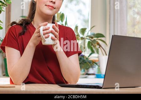 Selbstisolierung. Eine junge schöne glückliche Kaukasierin hält einen Becher in der Hand.auf dem Tisch ist ein Laptop. Konzept der Quarantäne, moderne Technologien a Stockfoto