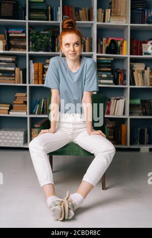 Glückliche Rothaarige junge Frau, die auf einem Stuhl im Bibliotheksraum vor dem Hintergrund von Bücherregalen sitzt. Stockfoto