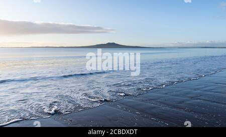 Milford Strand bei Sonnenaufgang mit wirbelnden Wellen im Vordergrund Und Rangitoto Island in der Ferne Stockfoto