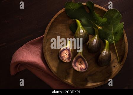 Saftig frische ganze Feigenfrüchte und eine in die Hälfte geschnitten Auf runder Olivenholzplatte auf dunklem Holzhintergrund mit abb.-Lamellen Stockfoto