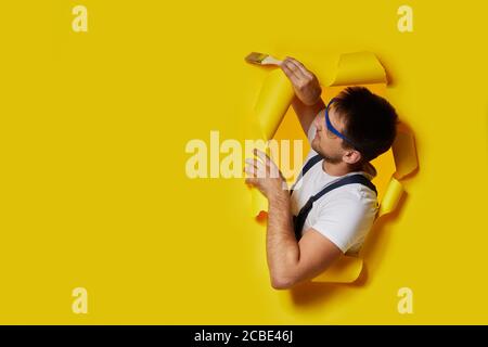 kaukasischen Maler Arbeiter in Sicherheitsglas und Uniform halten Pinsel durch Loch in gelbem Papier. Handwerker Malerei Wand. Kopieren Raum Stockfoto