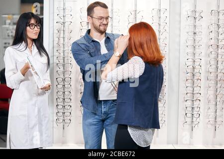 Ginger Frau versucht auf einem neuen Paar Brille, ihr Freund hilft ihr, während asiatische schöne Optiker hält einen Spiegel. Nahaufnahme Foto Stockfoto