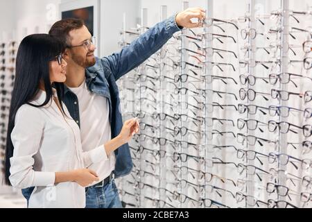 Bärtige handsoem lächelnden Mann Gläser aus dem Regal zu nehmen, um sie auszuprobieren, Nahaufnahme, Seite, Ansicht Foto. Kopieren Sie Platz Stockfoto