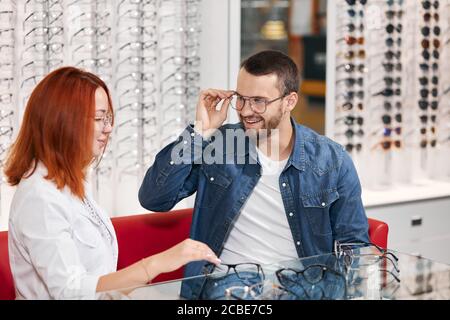 Junge positiver Mann in Jeans Jacke versuchen auf verschiedenen viele Gläser, bis Foto schliessen. Gläser auf dem Regal im Hintergrund des Fotos Stockfoto