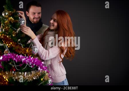Charmante komfortabel gekleidete Frau Dekoration Weihnachtsbaum zusammen mit positiven Freund, posiert über dunkelgrauen Hintergrund, Zeit, um neu zu feiern Stockfoto