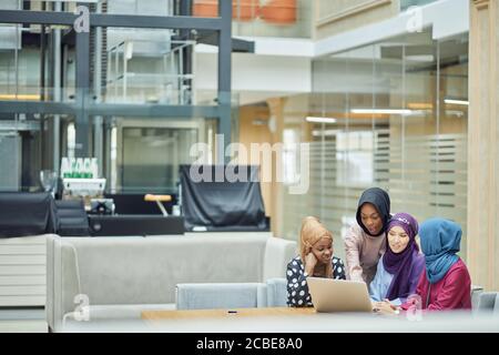 Glückliche junge muslimische asiatische und afrikanische Frauen in Kopfschals Und Hijab mit Laptop im Café mit Freunden Stockfoto