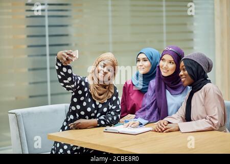 Vier asiatische muslimische Studenten Mädchen sitzen zusammen, tragen Hijabs und lange traditionelle tragen Spaß nach dem Unterricht und Selfie in College-Klasse Stockfoto