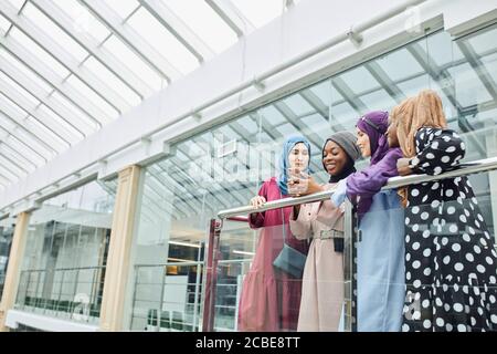 Gruppe von islamischen Frauen unterschiedlicher ethnischer Herkunft in smart gekleidet Traditionelle Kleidung reden und beobachten am Telefon zusammen, während In der Nähe der Hand stehen Stockfoto