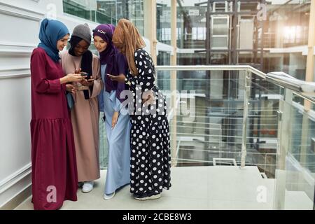 Arabian jung modisch in muslimischer Kleidung gekleidet Frauen beobachten auf Handy musikalischen Videoclip stehend zusammen im Einkaufszentrum - Millennials, ge Stockfoto