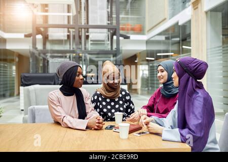 Muslimische junge Frauen unterschiedlicher Ethnie, in bunten nationalen Kleidung chillen im Café gekleidet, reden über neues Buch des berühmten Autors Stockfoto