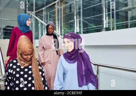 Eine Gruppe lächelnder muslimischer Frauen im Hijab, die die Treppe des Geschäftszentrums hinunter gingen. Arabische und afrikanische Damen in langen eleganten Kleidern und Schals gekleidet Stockfoto