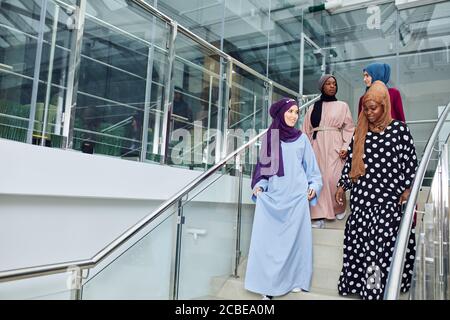 Junge muslimische, fröhliche Geschäftsreisende Frauen in nationalen arabischen Kleidern besuchen das Business Center, steigen die Treppe hinauf und unterhalten sich über ihre Fut Stockfoto