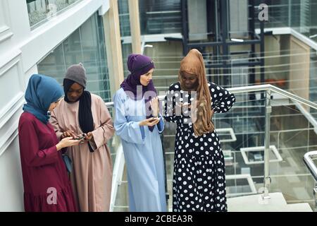 Arabian jung modisch in muslimischer Kleidung gekleidet Frauen beobachten auf Handy musikalischen Videoclip stehend zusammen im Einkaufszentrum - Millennials, ge Stockfoto