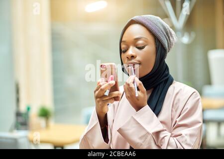 Schöne junge afrikanische muslimische Frau, die Lippen mit Lippenstift suchen Im Spiegel ihres Smartphones Stockfoto