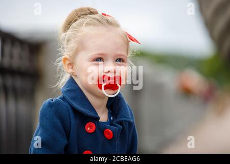 Kleines Mädchen in einem blauen Mantel mit einem Schnuller in Ihr Mund auf der Straße Stockfoto