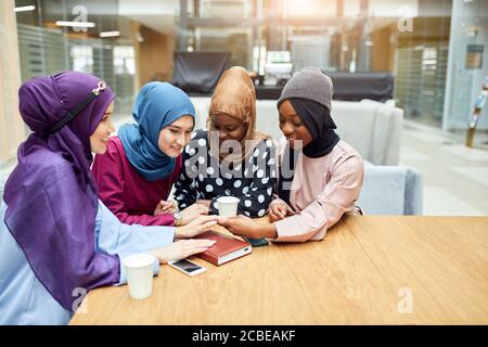 Porträt von smart elegant gekleidet kaukasischen und afrikanischen muslimischen weiblich Freunde, die die Hände einer der Frauen betrachten, die die Maniküre bewundern Während des Sitzens in CI Stockfoto