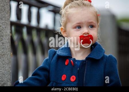 Kleines Mädchen in einem blauen Mantel mit einem Schnuller in Ihr Mund auf der Straße Stockfoto