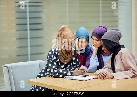 Islamische Reisebloggerin, die Informationen vom Smartphone mit ihr teilt Anhänger, während sie in der vierköpfigen Gruppe in der Hotellobby sitzen Stockfoto