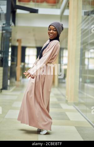 Modische fröhliche junge afrikanische muslimische Frau trägt rosa langes Kleid und Strickmütze auf schwarzem Schal, urbane Szene Stockfoto
