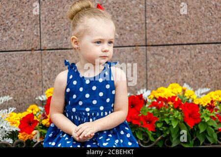 Ein kleines Mädchen in einem blauen Kleid und einem roten Bow sitzt mit farbigen Taschen auf der Straße daneben Blumen Stockfoto