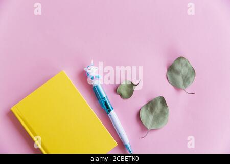 Gelbes Notizbuch, lustiger Einhorn-Stift und grüne Eukalyptusblätter auf rosa Pastellhintergrund. Flach liegend. Draufsicht. Speicherplatz kopieren Stockfoto