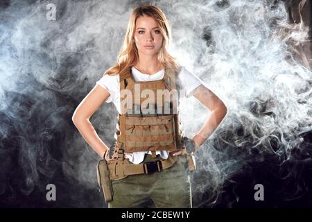 Portrait schöne blonde weibliche Soldat in Platte Träger und Gürtel gekleidet. Frauen, Armee, Waffe, Technologie und Menschen Konzept. Schwarze rauchige Hinterbeine Stockfoto