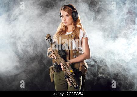 Schöne kaukasische weibliche private militärische Auftragnehmer mit langen blonden Haaren, gekleidet in taktische Uniform, bewaffnet mit Riffel posiert isoliert im Studio Stockfoto