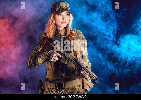 Soldatin in militärischer Tarnuniform und Mütze mit Scharfschütze Gewehr auf schwarzem Hintergrund mit rauchigen Wolken Stockfoto