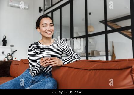 Attraktive lächelnde junge asiatische Frau trägt Freizeitkleidung Entspannung auf einer Ledercouch zu Hause, mit Handy Stockfoto