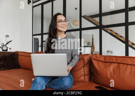 Attraktive lächelnde junge asiatische Geschäftsfrau entspannen auf einer Ledercouch zu Hause, arbeiten auf Laptop-Computer Stockfoto