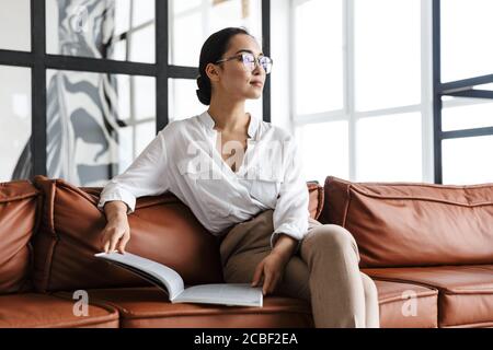 Attraktive lächelnde junge asiatische Geschäftsfrau, die sich zu Hause auf einer Ledercouch entspannt und ein Magazin liest Stockfoto