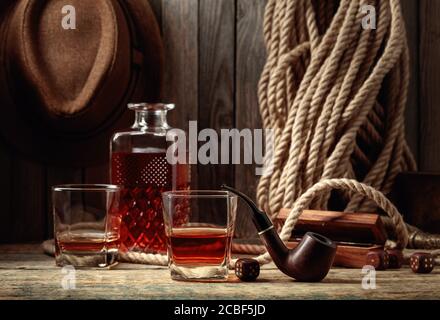 Glas und Dekanter mit einem starken alkoholischen Getränk auf einem alten Holztisch. Im Hintergrund sind ein Hanfseil und ein Männerhut. Stockfoto