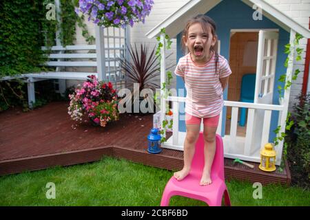 Das vierjährige, weiße Mädchen stand auf einem pinken Kinderstuhl aus Plastik und schrie vor einem blau-weißen Wendy-Haus. England GB Stockfoto