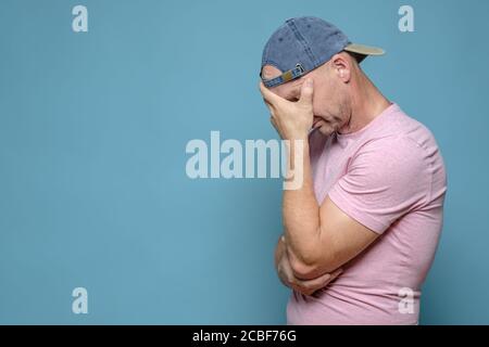 Mann in der Mütze und T-Shirt war sehr müde, aufgeregt und gestresst, bedecktes Gesicht mit Hand und gesenktem Kopf. Speicherplatz kopieren. Blauer Hintergrund. Stockfoto