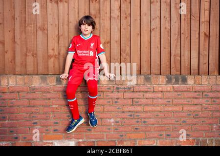 Der neunjährige kaukasische Junge sitzt an einer Außenwand und trägt das 2020/21 Heim Liverpool Fußballtrikot, das von Nike entworfen wurde. Lancashire UK Stockfoto