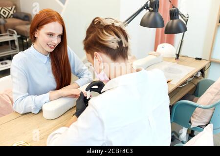 Manikurist Feilen und Formen Nägel während der Prozedur der Maniküre im Schönheitssalon. Rotschopf Frau lächeln und genießen Prozess. Schönheitskonzept Stockfoto