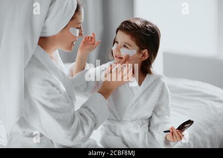 Beauty-Konzept. Sorgfältige aufmerksame Mutter verwenden Flecken für Augen und ihre Tochter in Maske zu. Eltern und Kinder interessiert an Schönheitsverfahren Stockfoto