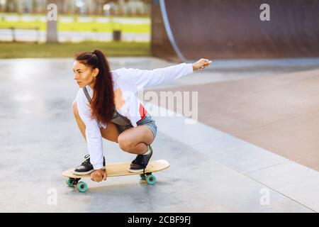 Braunhaarige Frau reitet im Park auf einem Skateboard. Model posiert auf dem Sportplatz Stockfoto