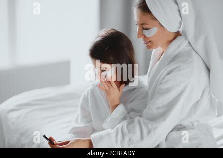 Seitenansicht auf schöne Mutter und Tochter tragen Bademantel und Handtücher verwenden Gesichtsmasken zu Hause, Flecken unter dem Auge. Kleines Kind hält Spiegel Stockfoto