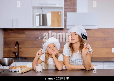 kaukasische Frau und Tochter denken über zukünftige Backen, Kochen in der Küche zusammen, tragen Schürze und Kappen zum Kochen Stockfoto