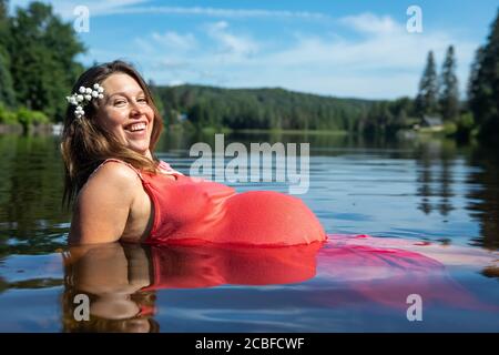 Schöne schwangere junge Frau in einem ärmellosen roten Kleid sitzt mit ihrem großen Bauch im Wasser eines kanadischen Sees und lächelt vor der Kamera. Stockfoto