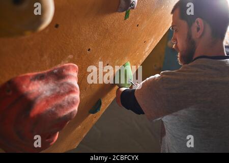 Nahaufnahme eines jungen, körperlich behinderten Klettertrainers, Inspektion und Fixierung von Griffen an einer künstlichen Wand in der Boulderhalle vor dem Klettern c Stockfoto