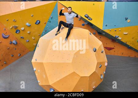 Junge fröhliche körperlich behinderte Boulderer sitzen auf der großen runden Form künstlichen Felswand, zeigt seine Bizeps, absolut glücklich und Stockfoto