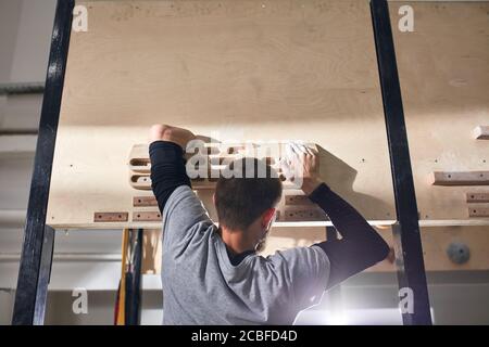 Nahaufnahme des jungen Kletterers ohne Unterarm, der an einer speziellen Holzplatte hängt, um festen Halt zu trainieren und in Kletterräumen zu trainieren. Stockfoto